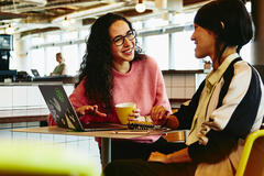 Two woman sitting in restaurant having a converstation, looking at laptop, smiling.