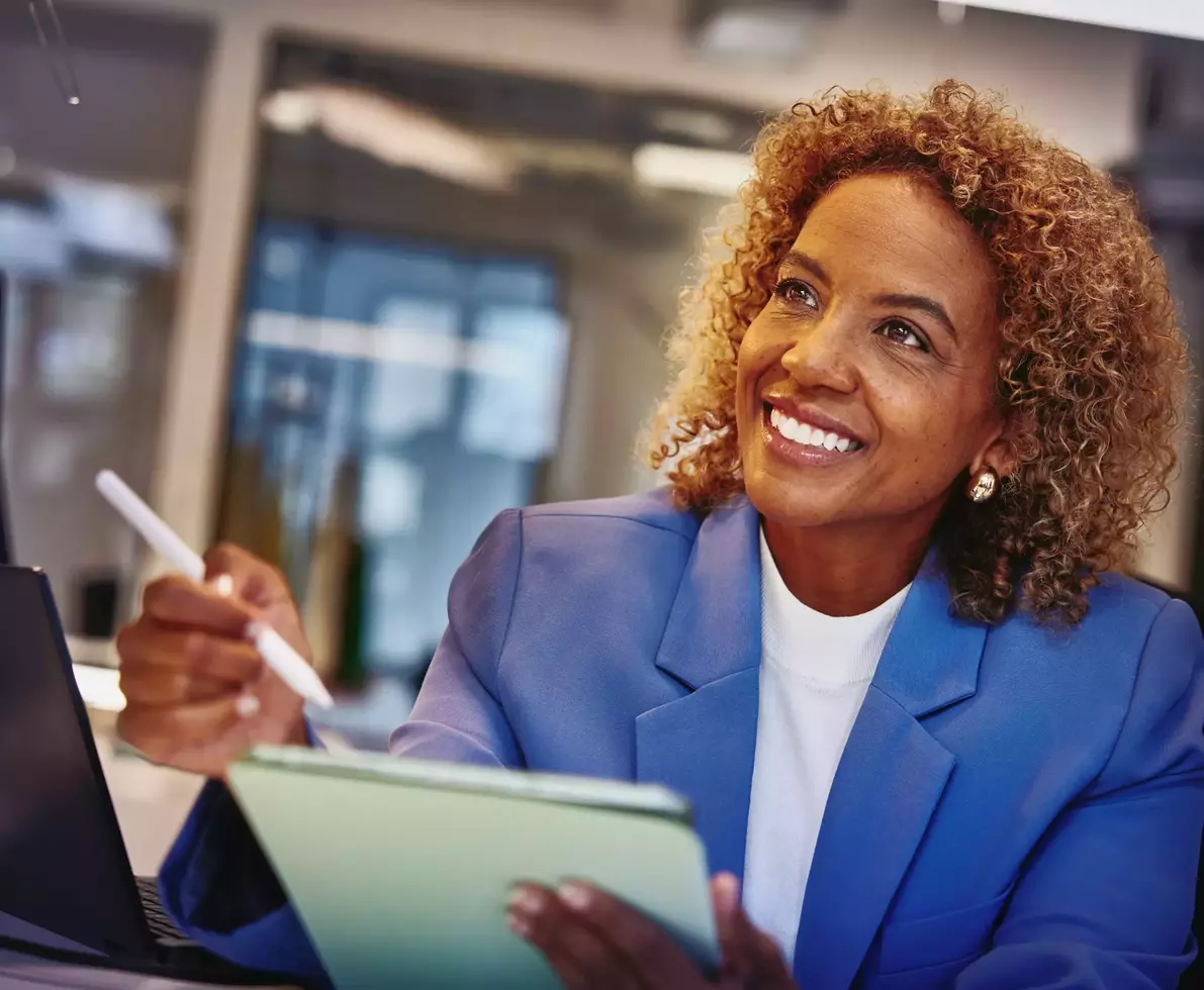 Female with blue blazer holding a touch pen and a tablet, sitting at a desk